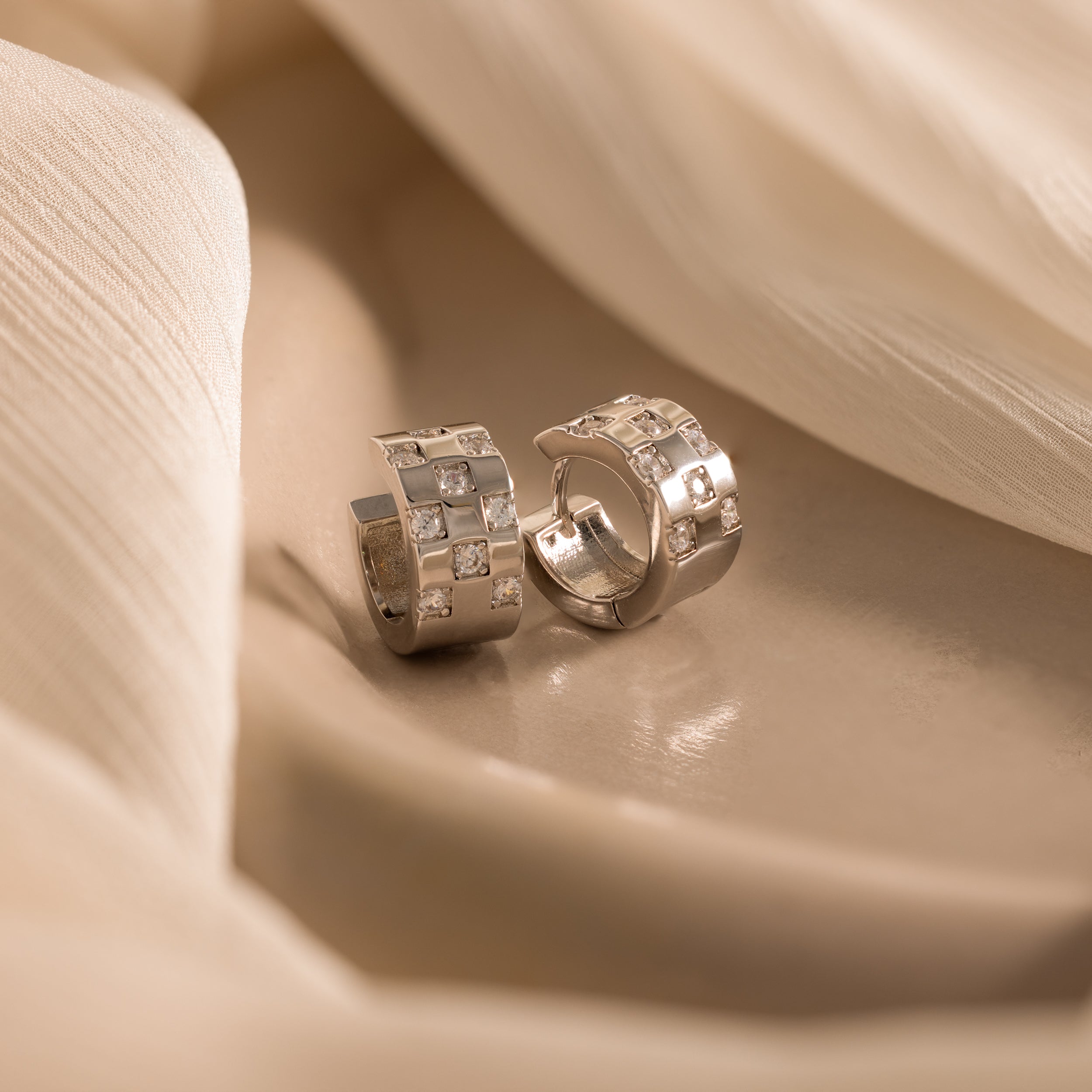 Two Pave Checkered Huggies in Silver with square gem details rest on a soft beige fabric background.