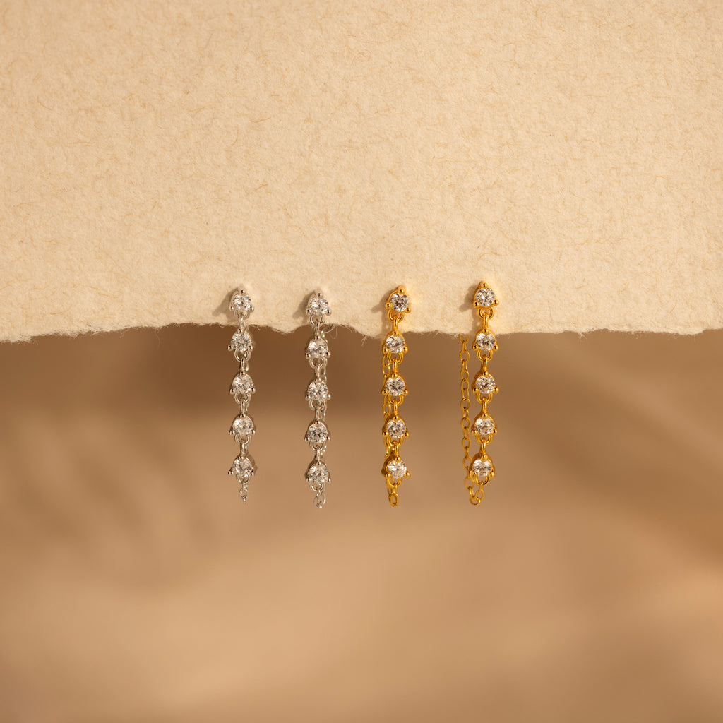 Two pairs of Diamond Chain Drop Earrings, one silver and one gold, are displayed on a beige textured background—add elegance to any occasion with these stunning earrings.