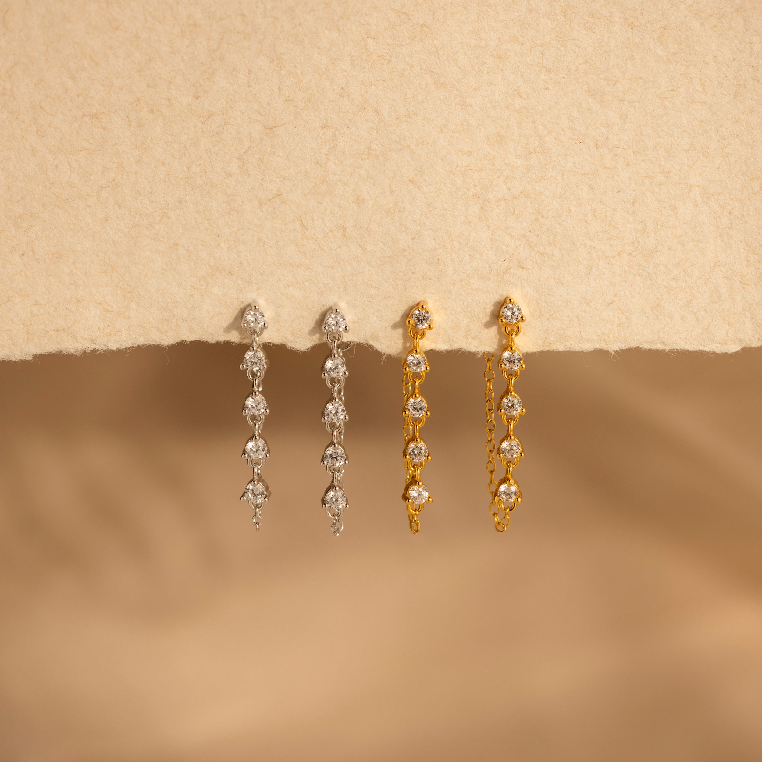 Two pairs of Diamond Chain Drop Earrings, one silver and one gold, are displayed on a beige textured background—add elegance to any occasion with these stunning earrings.