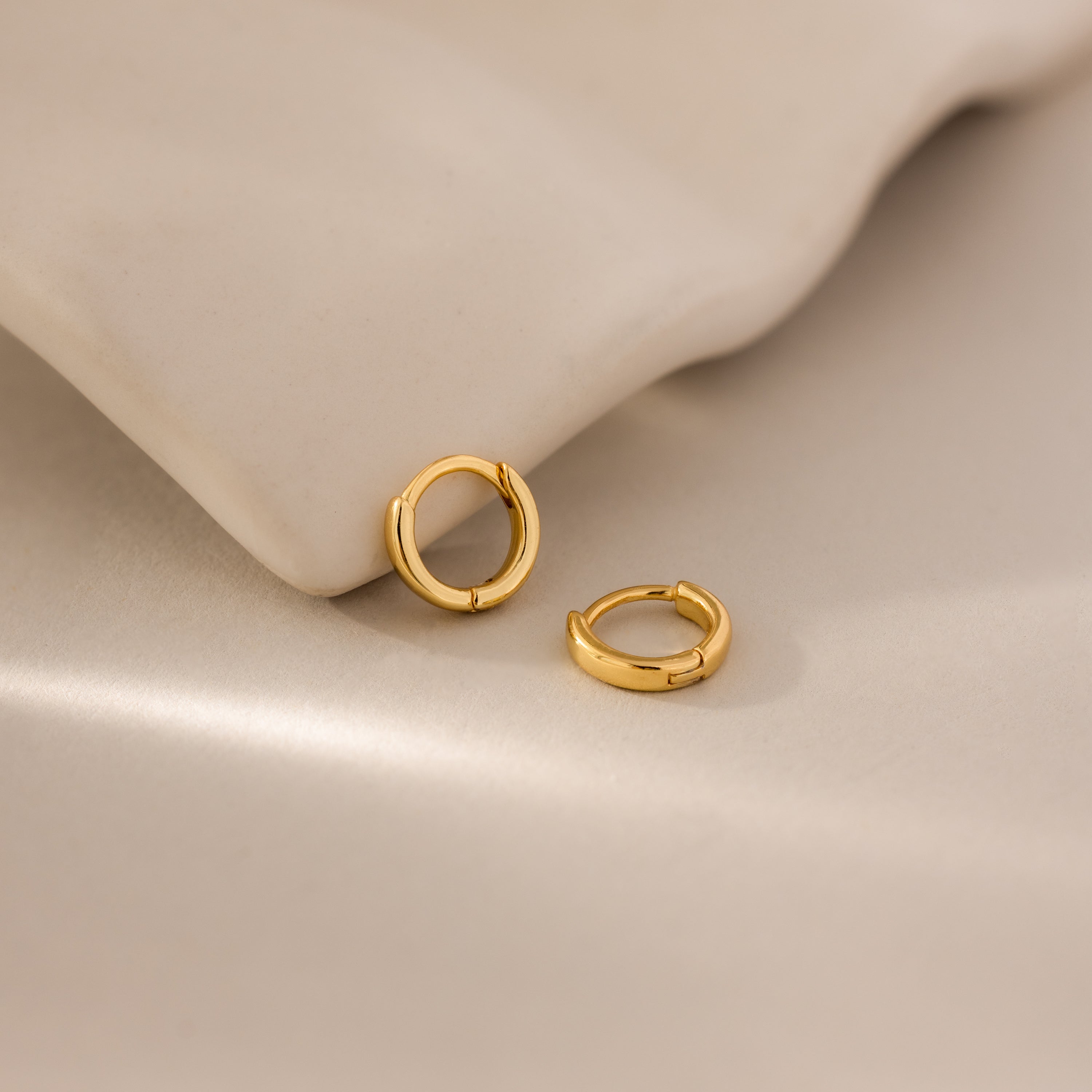 Pair of 6mm tiny classic gold huggie hoop earrings resting on a neutral surface, styled beside a smooth ceramic dish.