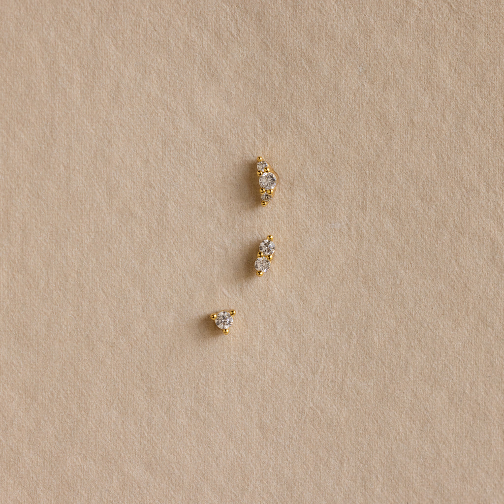 Three Tiny Diamond Flatback Studs Set in 18K Gold are arranged vertically on a textured beige surface.