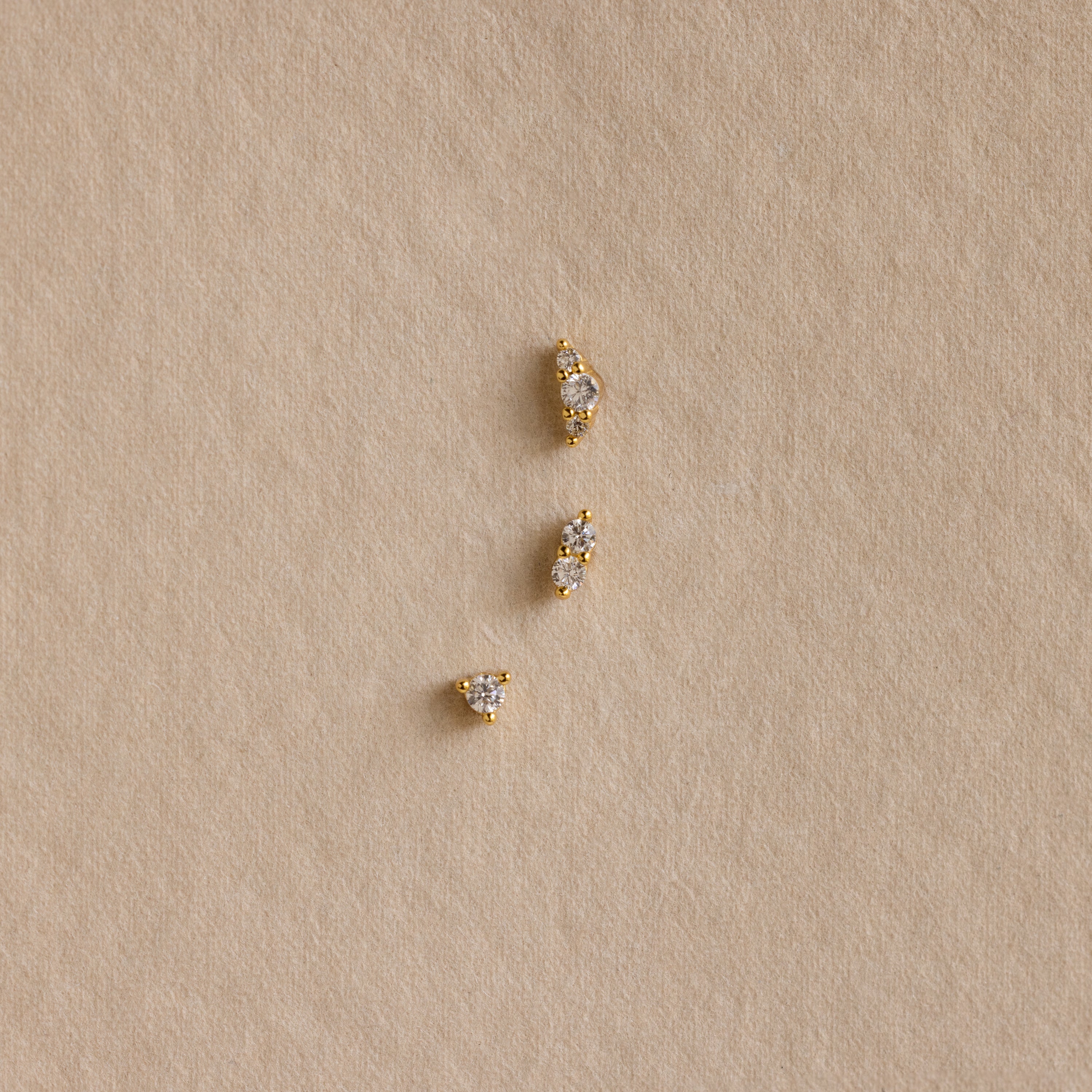 Three Tiny Diamond Flatback Studs Set in 18K Gold are arranged vertically on a textured beige surface.