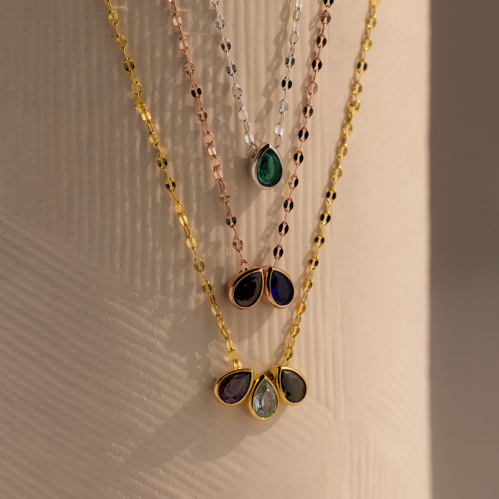 The Multiple Teardrop Birthstone Necklace features four layered, colorful gemstone pendants—including a teardrop birthstone—displayed on a textured beige surface, making it a perfect personalized keepsake jewelry piece.
