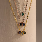The Multiple Teardrop Birthstone Necklace features four layered, colorful gemstone pendants—including a teardrop birthstone—displayed on a textured beige surface, making it a perfect personalized keepsake jewelry piece.