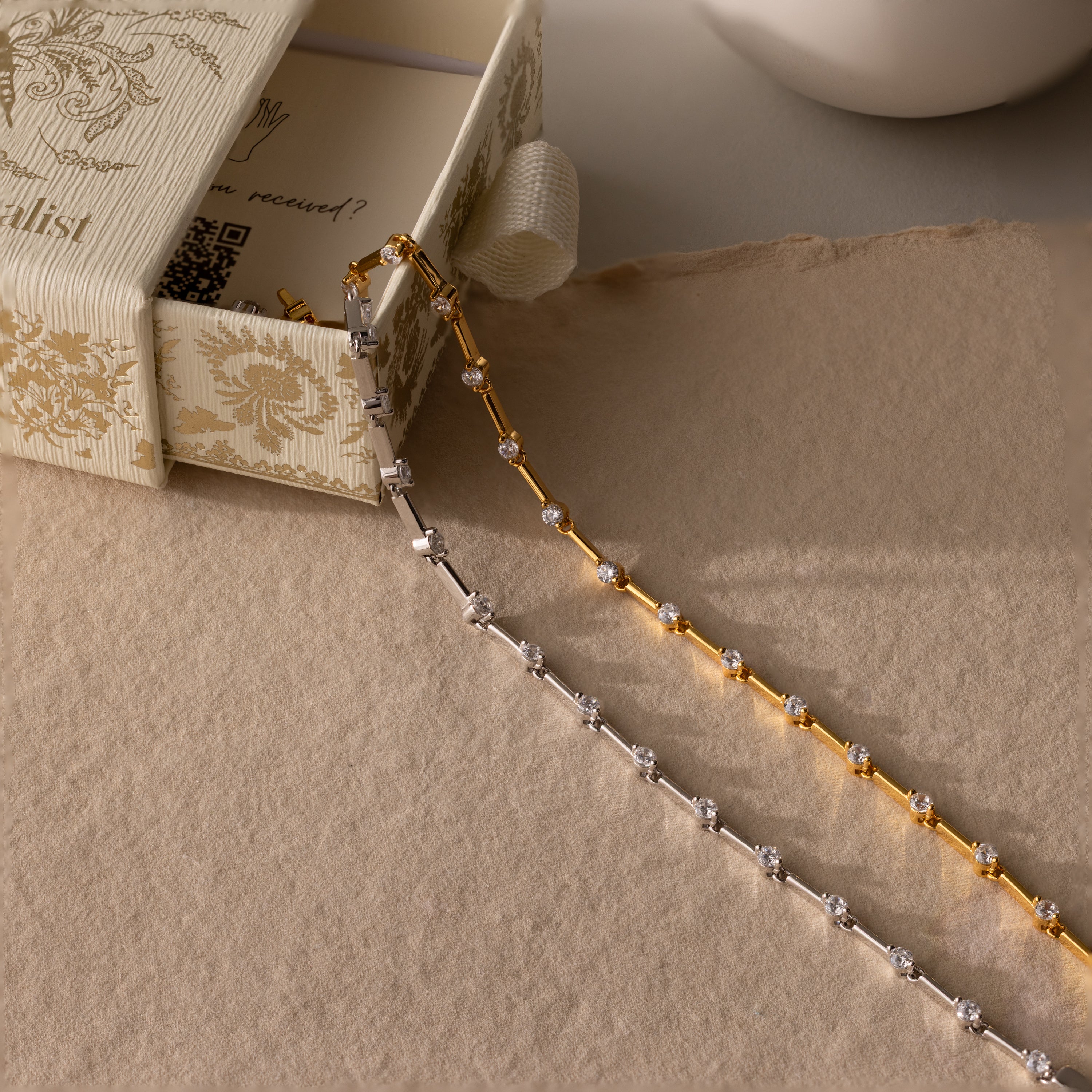 Two delicate chain necklaces—one gold, one silver—shown beside an ornate cream box, pair perfectly with the Sprinkle Diamond Tennis Bracelet in 18K Gold.