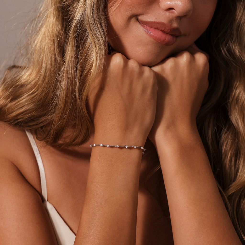 A woman with wavy hair rests her chin on her hands, wearing a cream top and the Sprinkle Diamond Tennis Bracelet, which adds an elegant touch to her look.