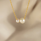 The Dainty Duo Pearl Necklace features two white pearls of varying sizes on a gold chain, perfect as elegant wedding jewelry, displayed against a soft beige blurred background.