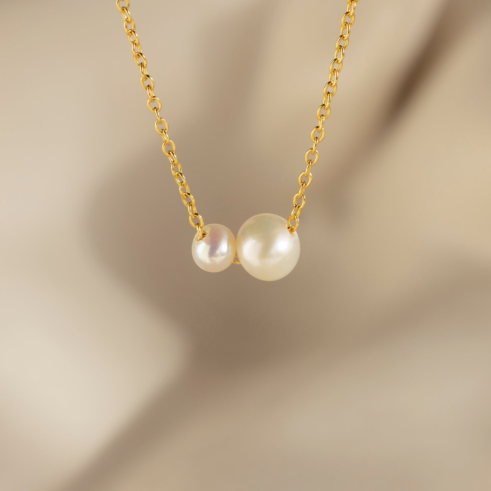 The Dainty Duo Pearl Necklace features two white pearls of varying sizes on a gold chain, perfect as elegant wedding jewelry, displayed against a soft beige blurred background.