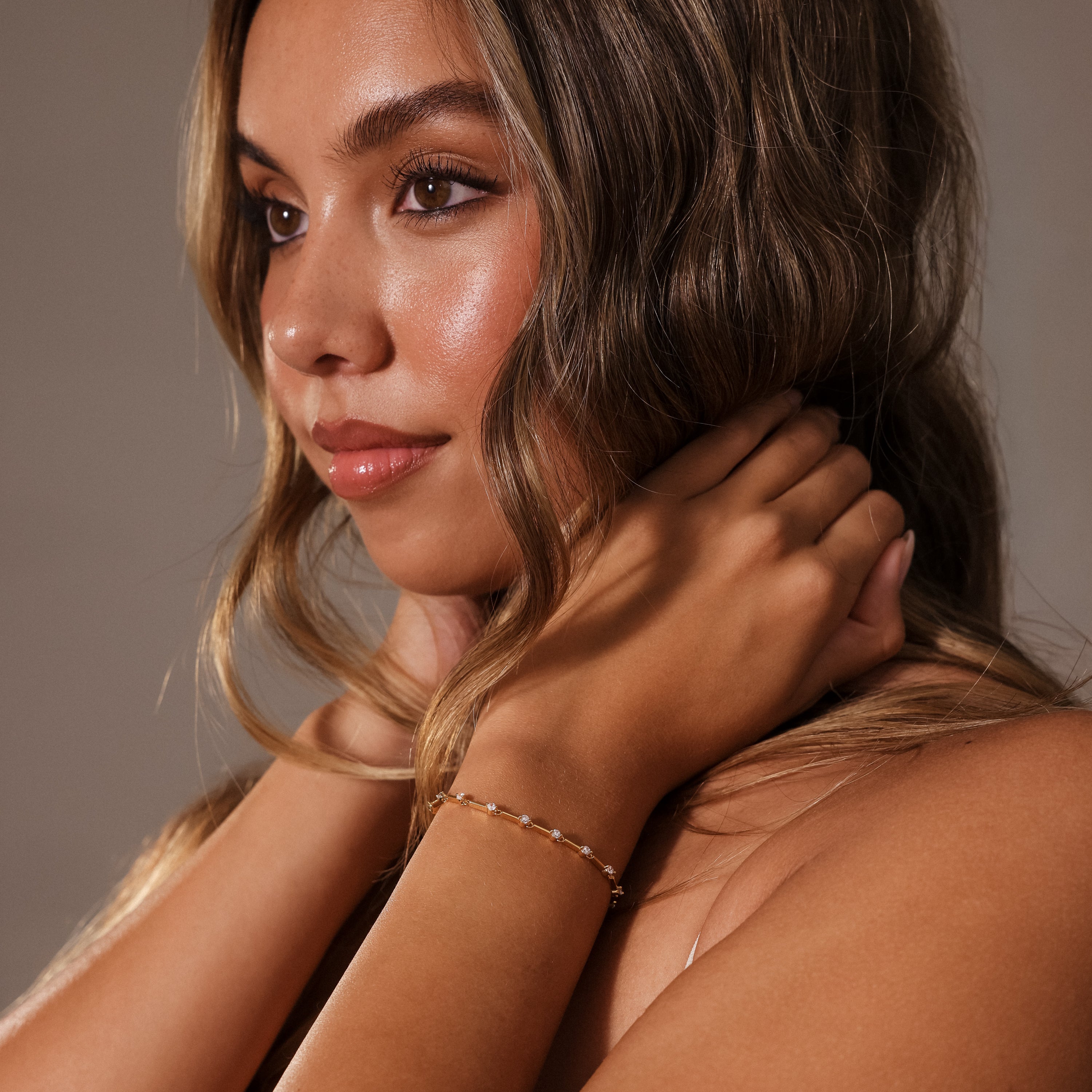 Woman with long wavy hair gazes to the side, hands near neck, wearing the Sprinkle Diamond Tennis Bracelet in 18K Gold.
