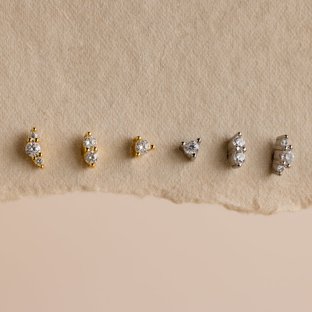 Six pairs from the Tiny Diamond Flatback Studs Set, in gold and silver, are arranged on a textured beige background, highlighting these elegant studs as the perfect accessory for any occasion.