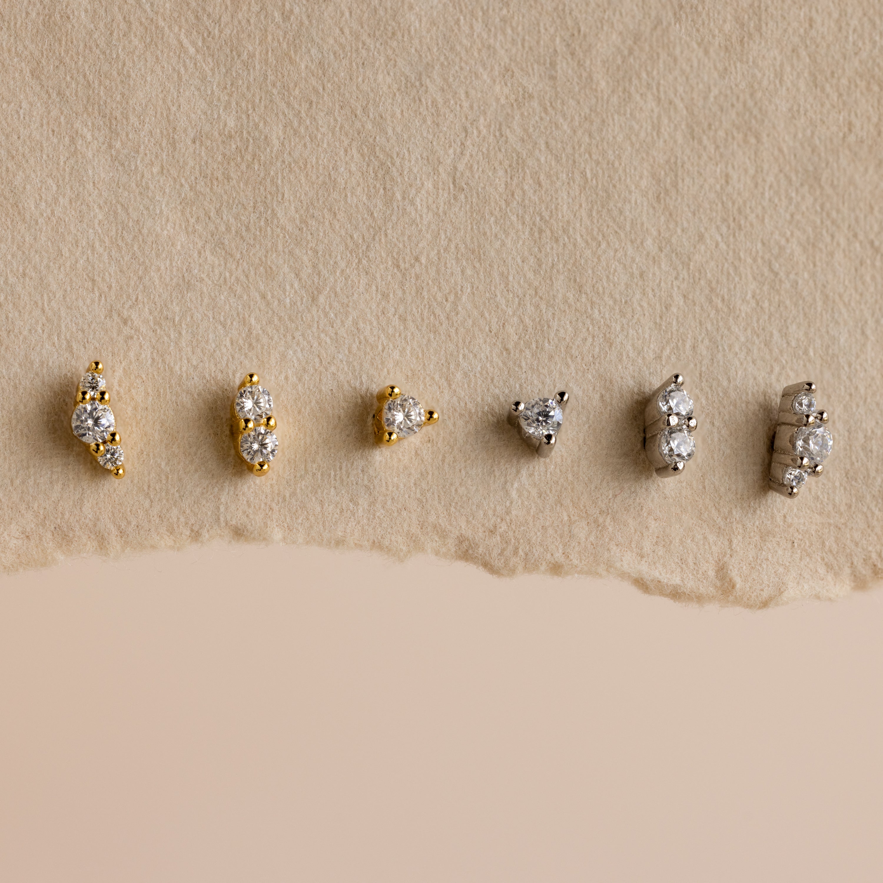 Six pairs from the Tiny Diamond Flatback Studs Set, in gold and silver, are arranged on a textured beige background, highlighting these elegant studs as the perfect accessory for any occasion.