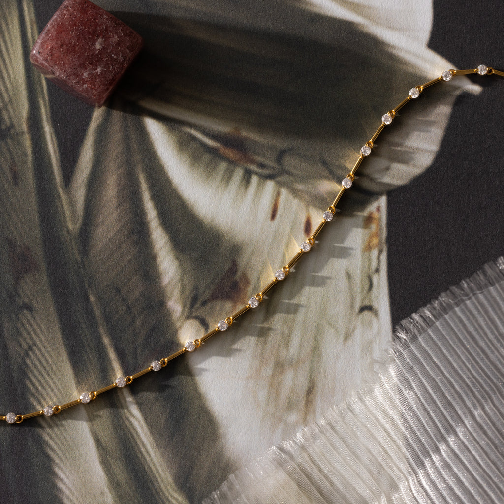The Sprinkle Diamond Tennis Bracelet in 18K Gold, displayed on patterned fabric beside a reddish stone and textured sheet, exudes delicate elegance and timeless charm.