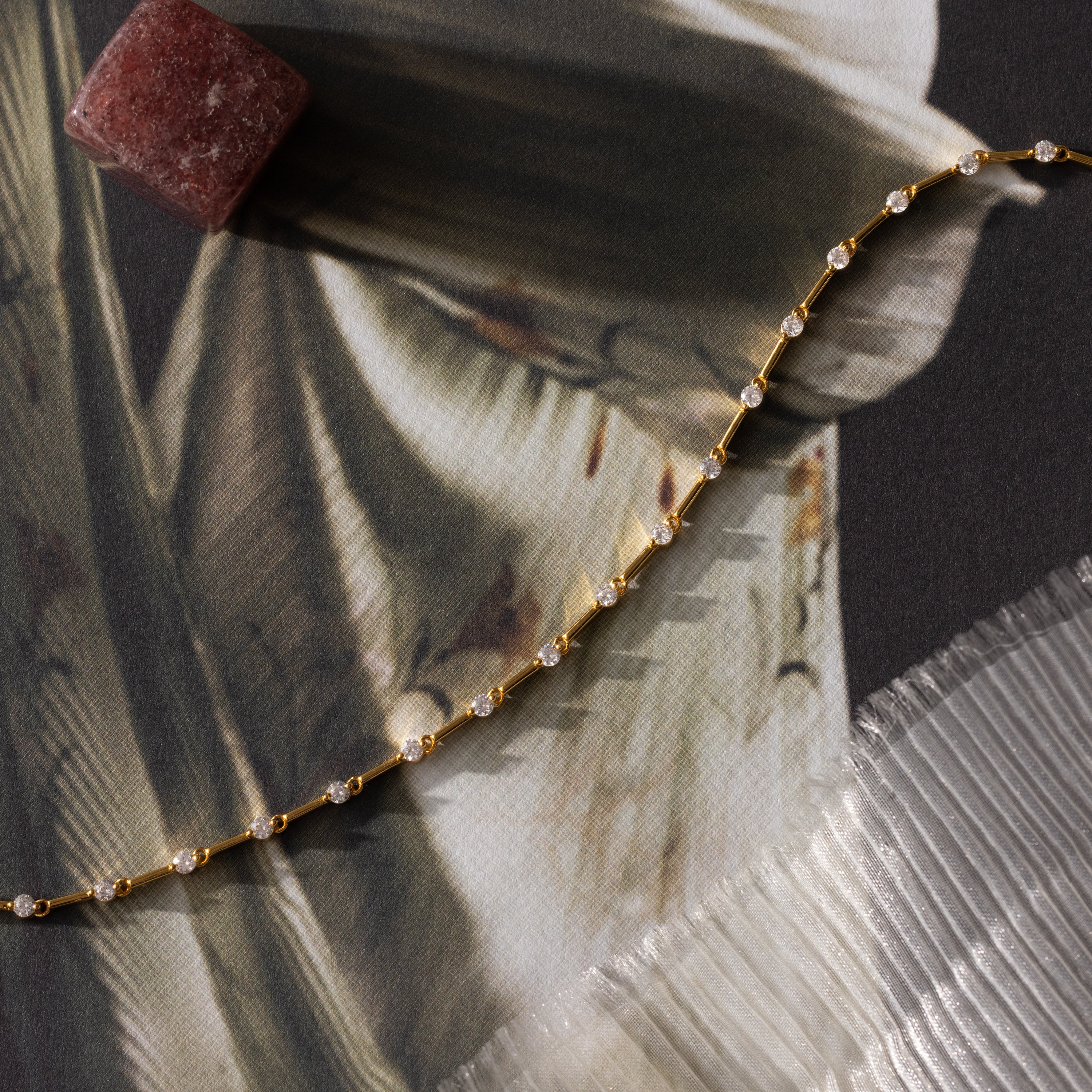 The Sprinkle Diamond Tennis Bracelet in 18K Gold, displayed on patterned fabric beside a reddish stone and textured sheet, exudes delicate elegance and timeless charm.