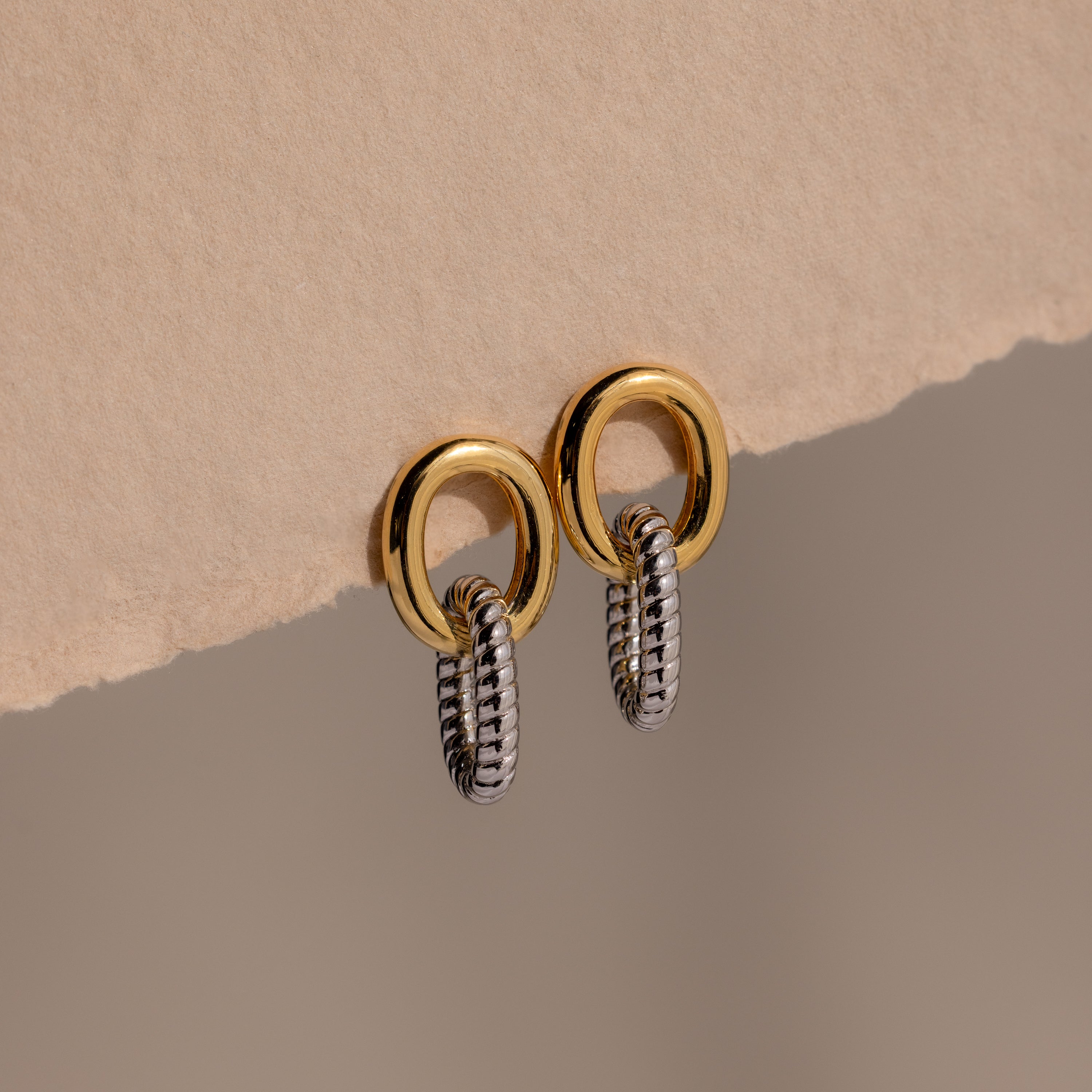 Twisted Link Earrings in Mixed Metal hang on a textured beige surface, displaying sculptural jewelry against a neutral background.