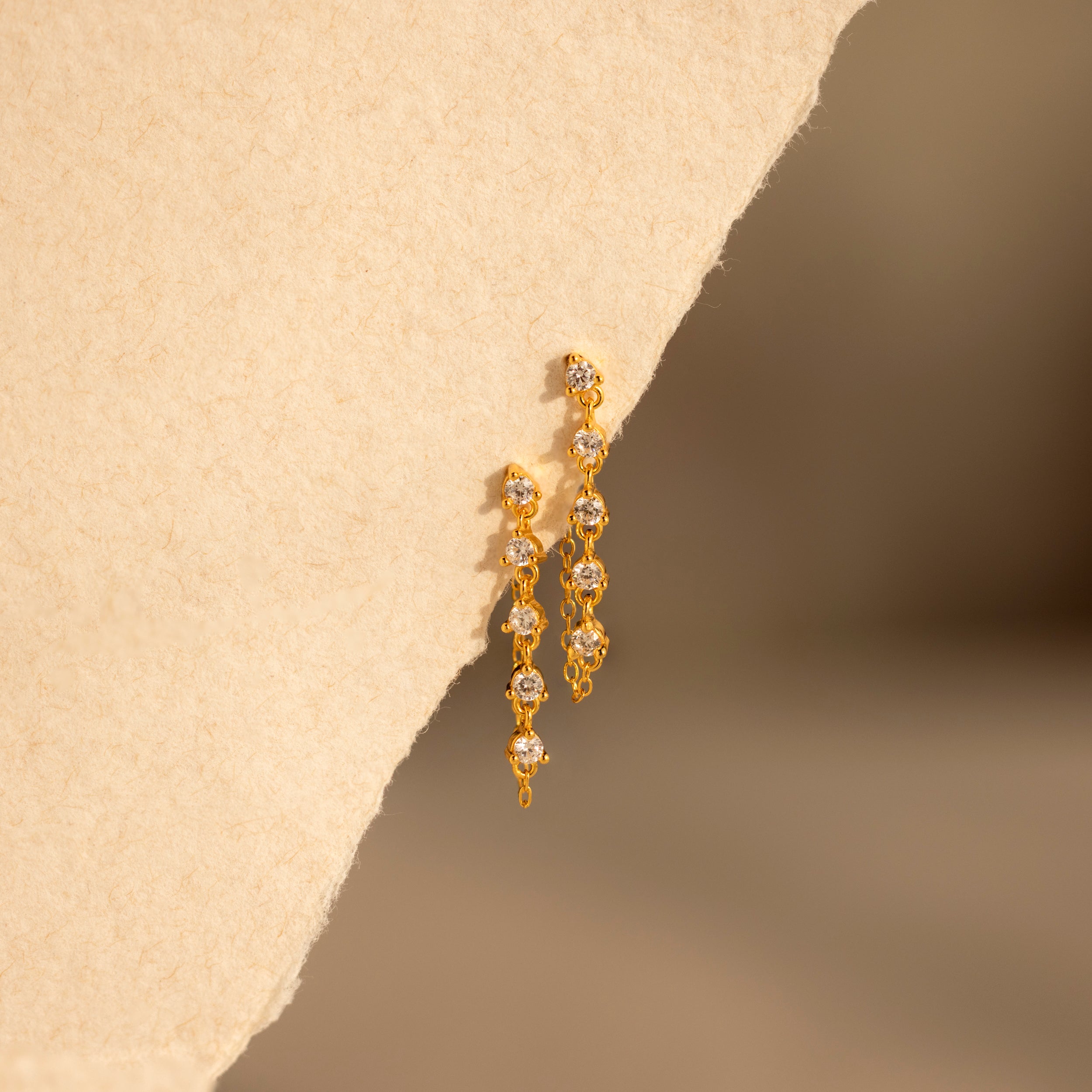 Diamond Chain Drop Earrings in gold feature small clear stones and are displayed on a textured beige surface with soft lighting—ideal for an elegant look.