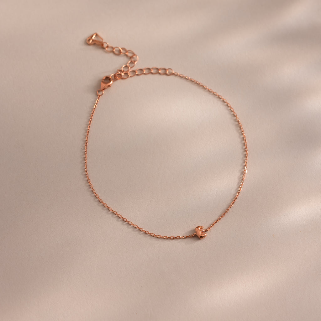Close up of a dainty rose gold bubble letter E bracelet against a neutral background.