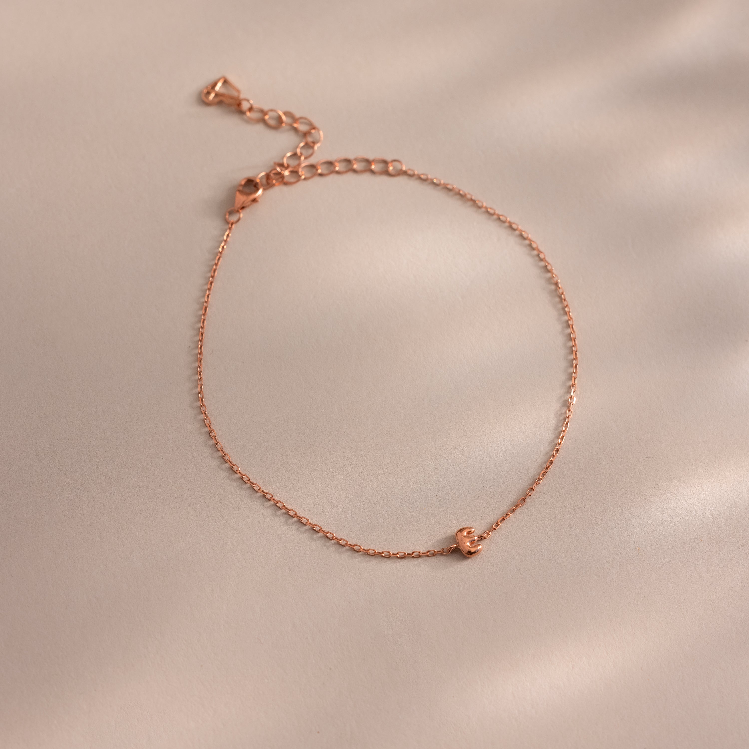 Close up of a dainty rose gold bubble letter E bracelet against a neutral background.