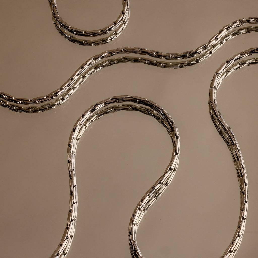 The Bold Link Chain Necklace in Silver features sleek, wavy links displayed on a beige reflective surface, capturing modern elegance and the striking appeal of an eye-catching statement necklace.
