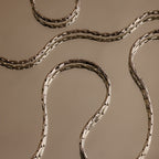 The Bold Link Chain Necklace in Silver features sleek, wavy links displayed on a beige reflective surface, capturing modern elegance and the striking appeal of an eye-catching statement necklace.