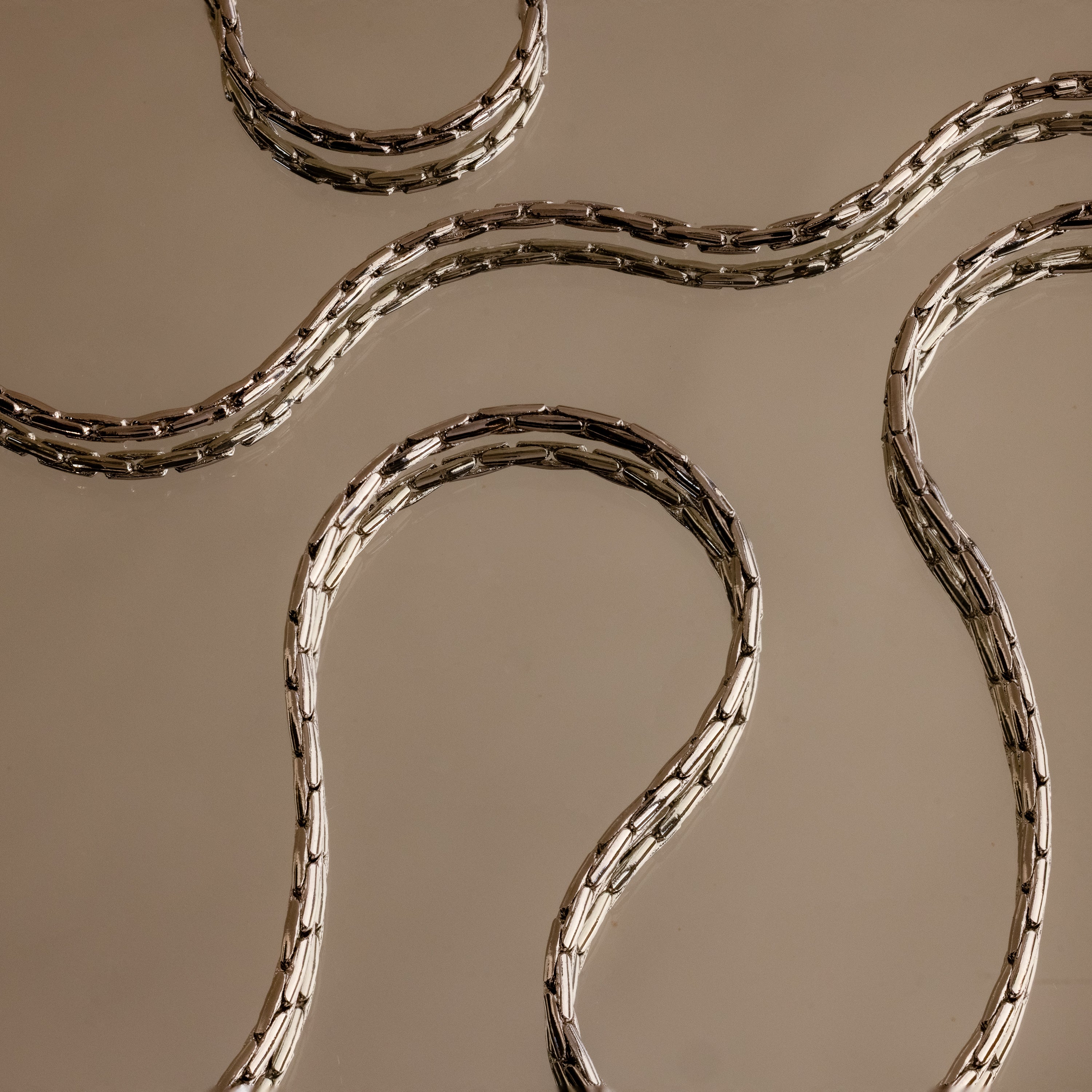 The Bold Link Chain Necklace in Silver features sleek, wavy links displayed on a beige reflective surface, capturing modern elegance and the striking appeal of an eye-catching statement necklace.