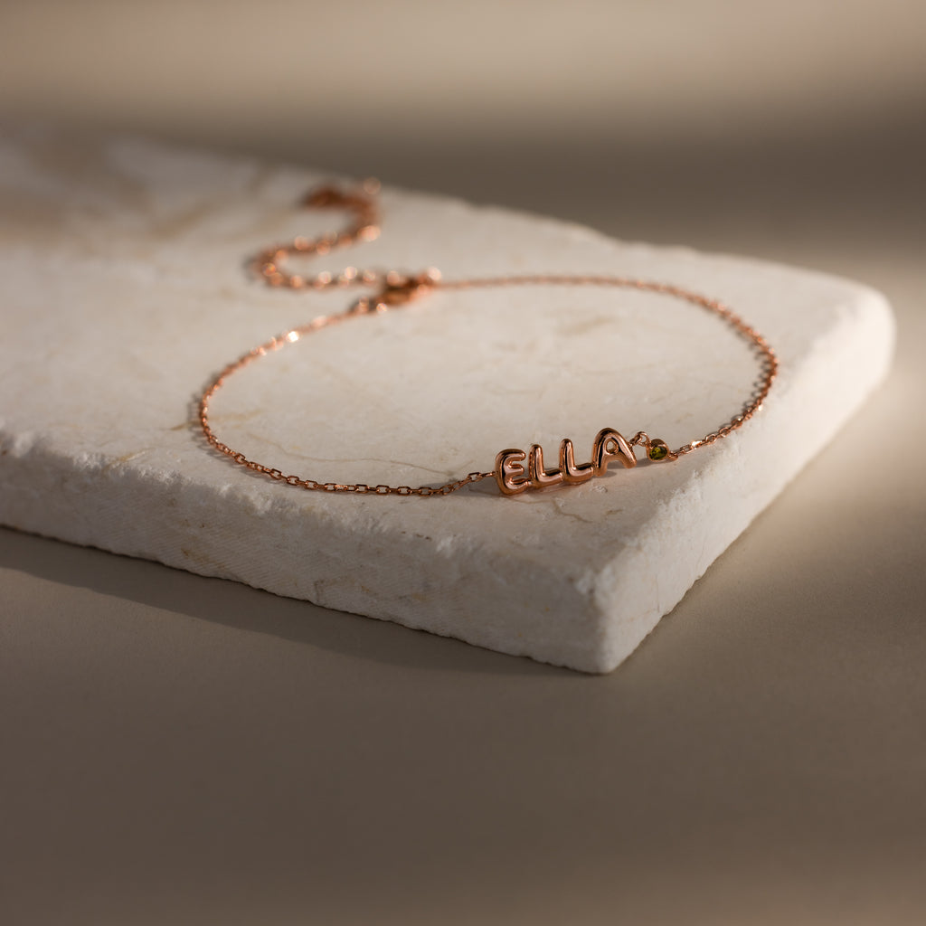 Rose gold dainty bubble letter bracelet with the name "ELLA" and a green peridot birthstone, displayed on a neutral surface.