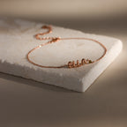 Rose gold dainty bubble letter bracelet with the name "ELLA" and a green peridot birthstone, displayed on a neutral surface.