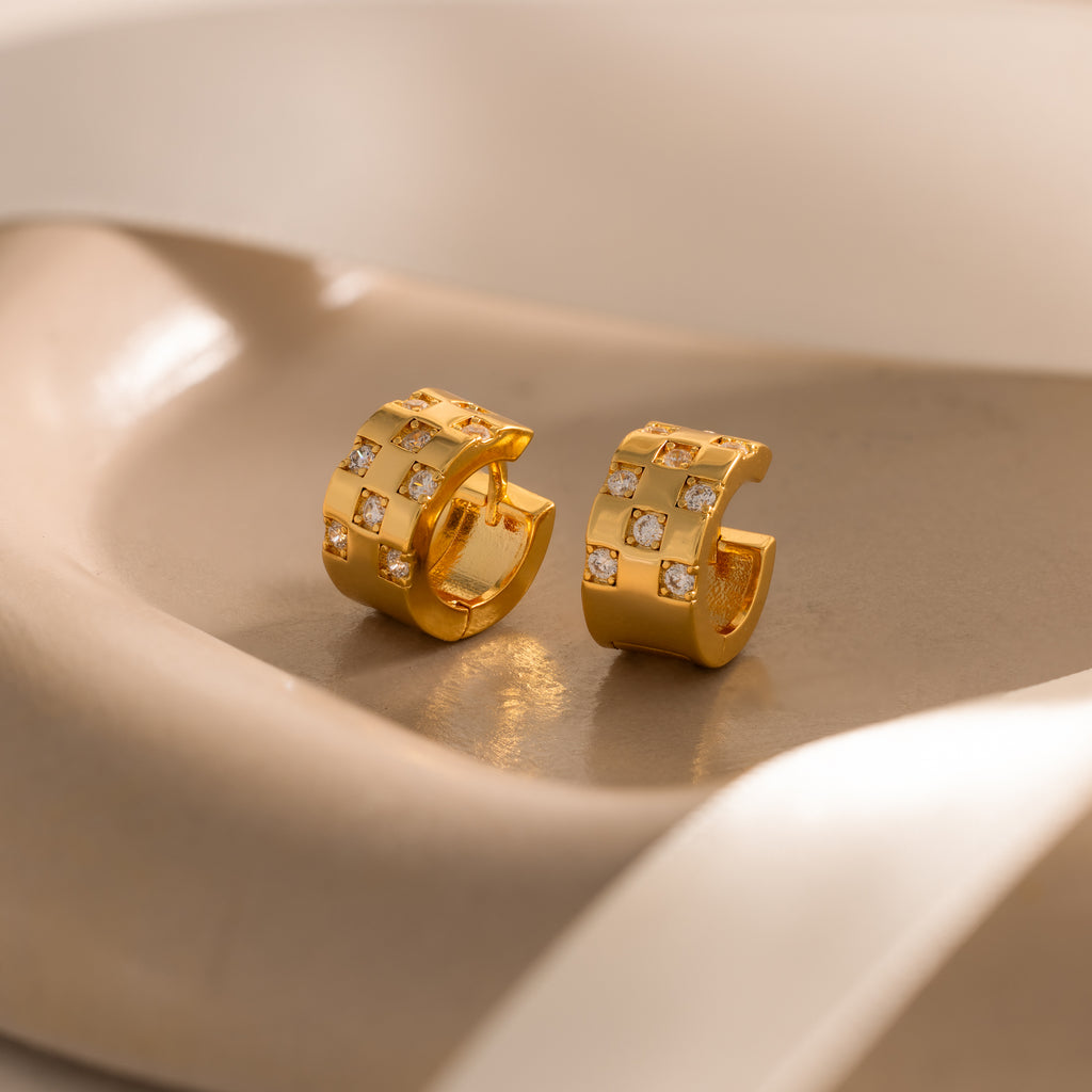 Pave Checkered Huggies in 18K Gold: gold chunky hoops with square patterns and embedded stones, displayed on a smooth beige surface.