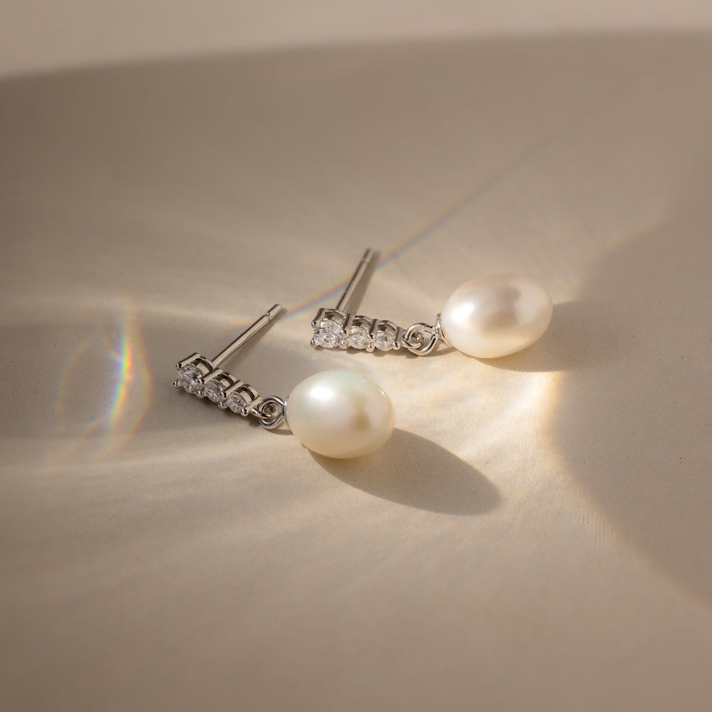 The Diamond Pearl Droplet Earrings rest on a beige surface, illuminated by soft sunlight and gentle shadows.