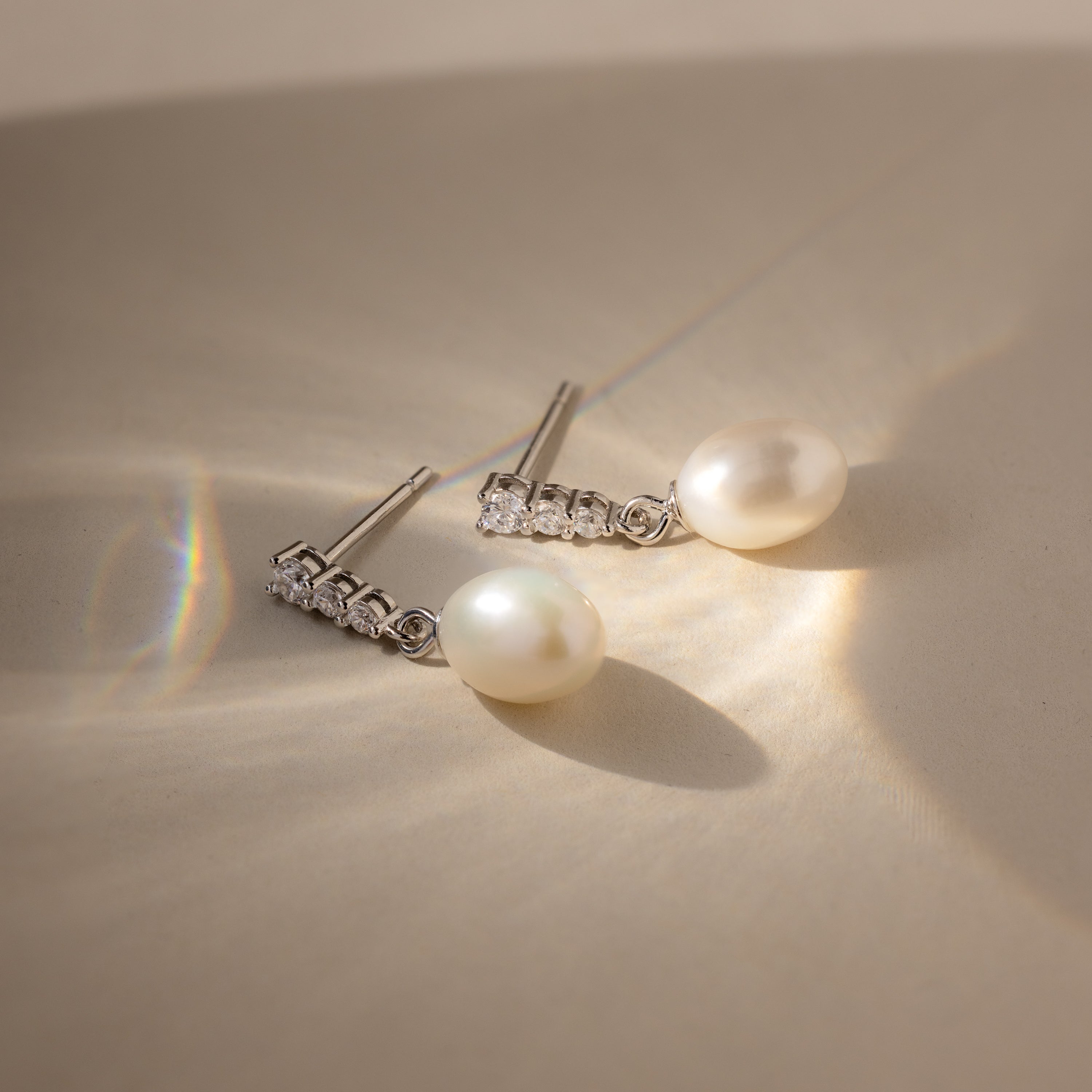 The Diamond Pearl Droplet Earrings rest on a beige surface, illuminated by soft sunlight and gentle shadows.