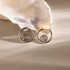 Spiral Swirl Hoops in Silver displayed before a white seashell on a beige background—ideal vacation jewelry or for giving your outfit a boho, beachy vibe.