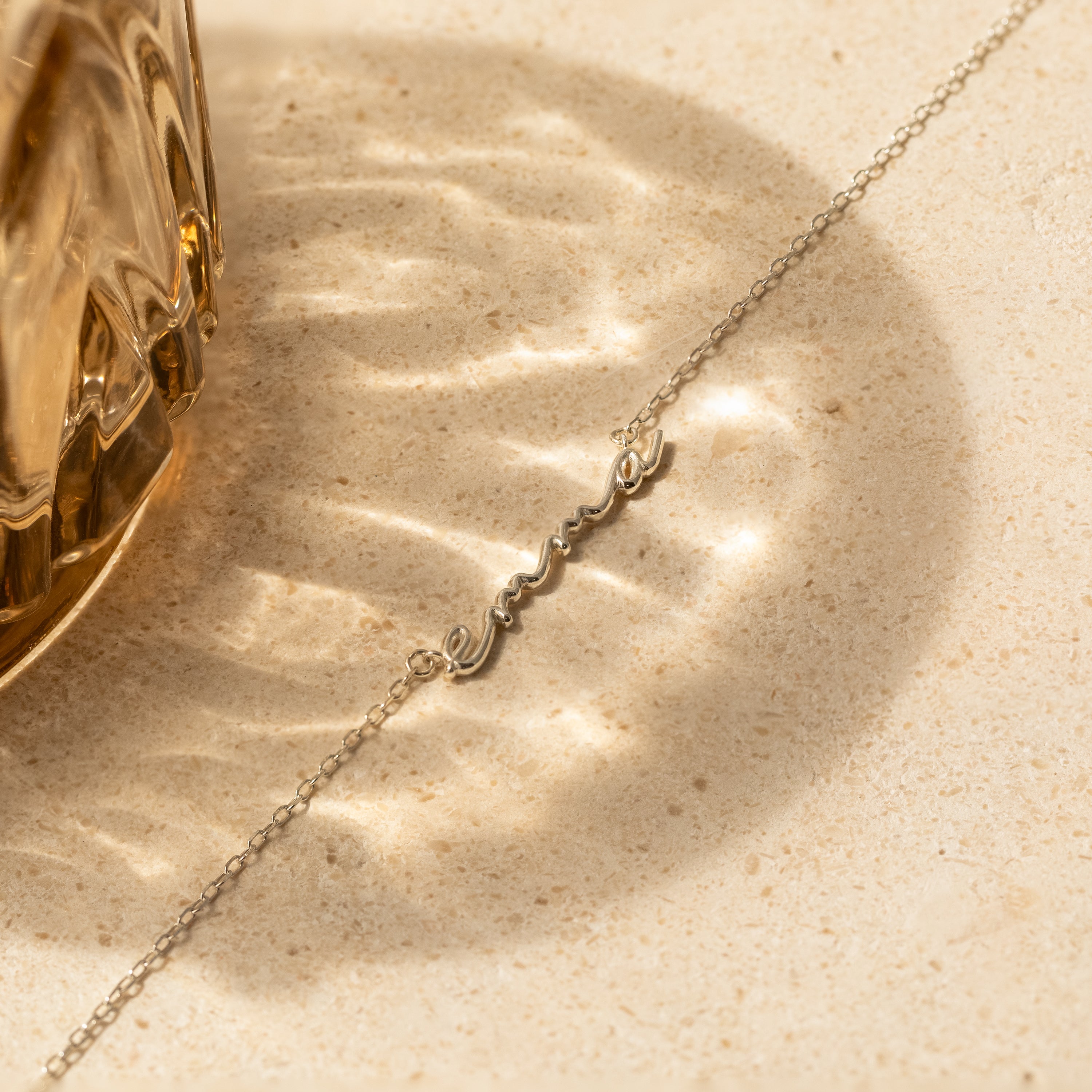 Dainty Bubble Name Necklace displayed on a beige stone surface with sunlight reflections, beside a glass object—perfect personalized nameplate for your custom jewelry collection.