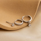 The Classic Diamond Earrings Set, featuring three elegant designs including a timeless stud, is displayed on a beige textured surface with a cream fabric background—ideal for wedding jewelry.