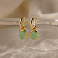 Jade Drop Hoops in 18K Gold with round jade gemstone accents hang from a clear rod on soft beige fabric, combining classic huggie style with vibrant elegance.