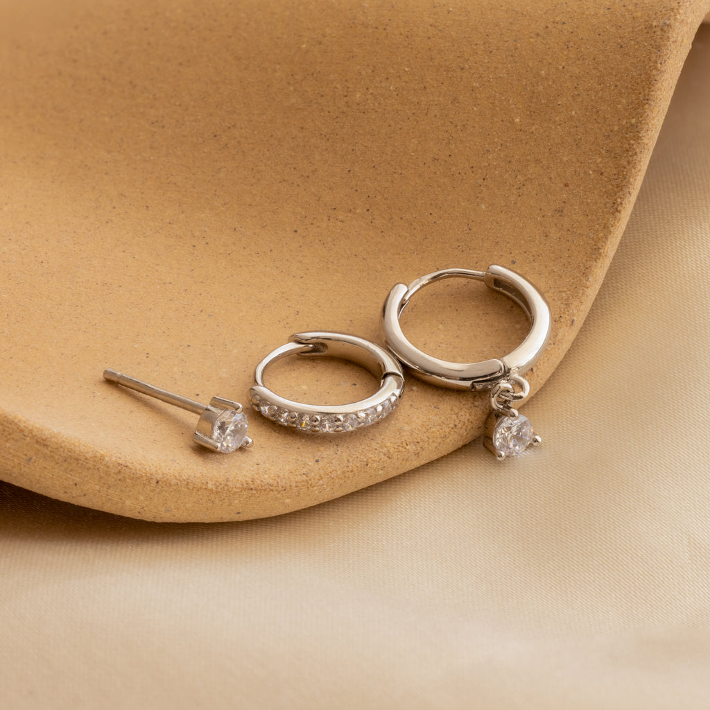 The Classic Diamond Earrings Set features three silver diamond earrings, including a classic stud design, beautifully displayed on tan and beige textured fabric—ideal for elegant wedding jewelry.