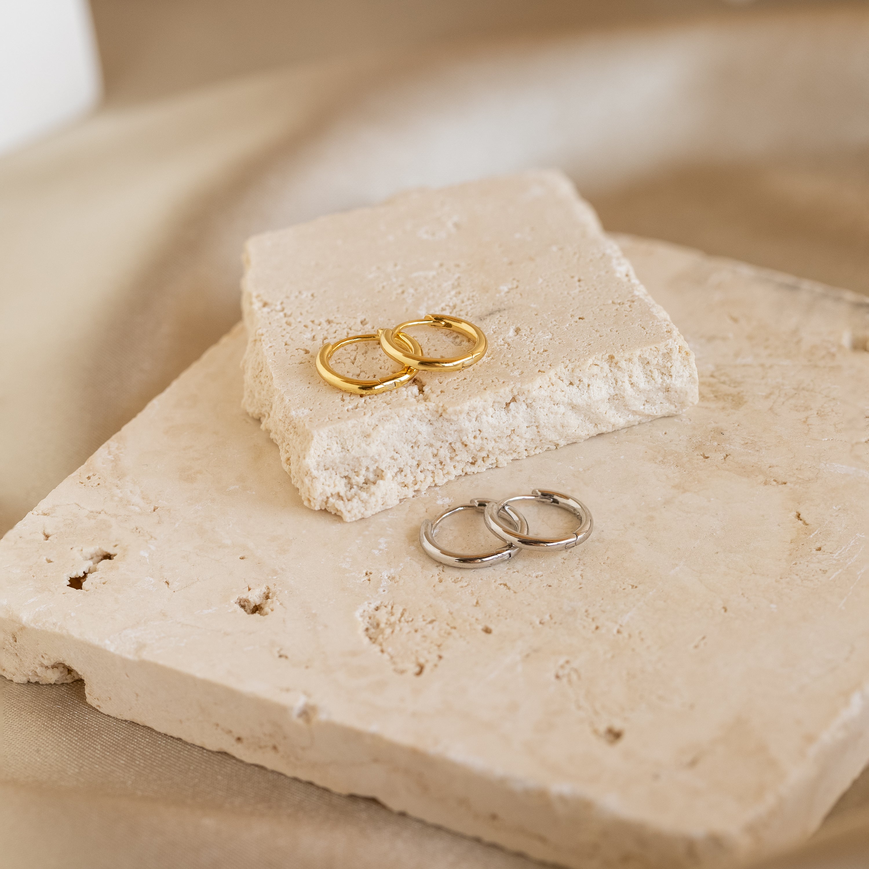 Two pairs of Small Bold Hoops, one gold and one silver, displayed on beige stone tiles—perfect minimalist earrings for everyday wear.