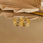 Two lightweight Medium Bold Hoops in 18K Gold rest on a reflective surface with an open book behind them. Perfect for everyday wear, these earrings bring a touch of elegance to your daily look.