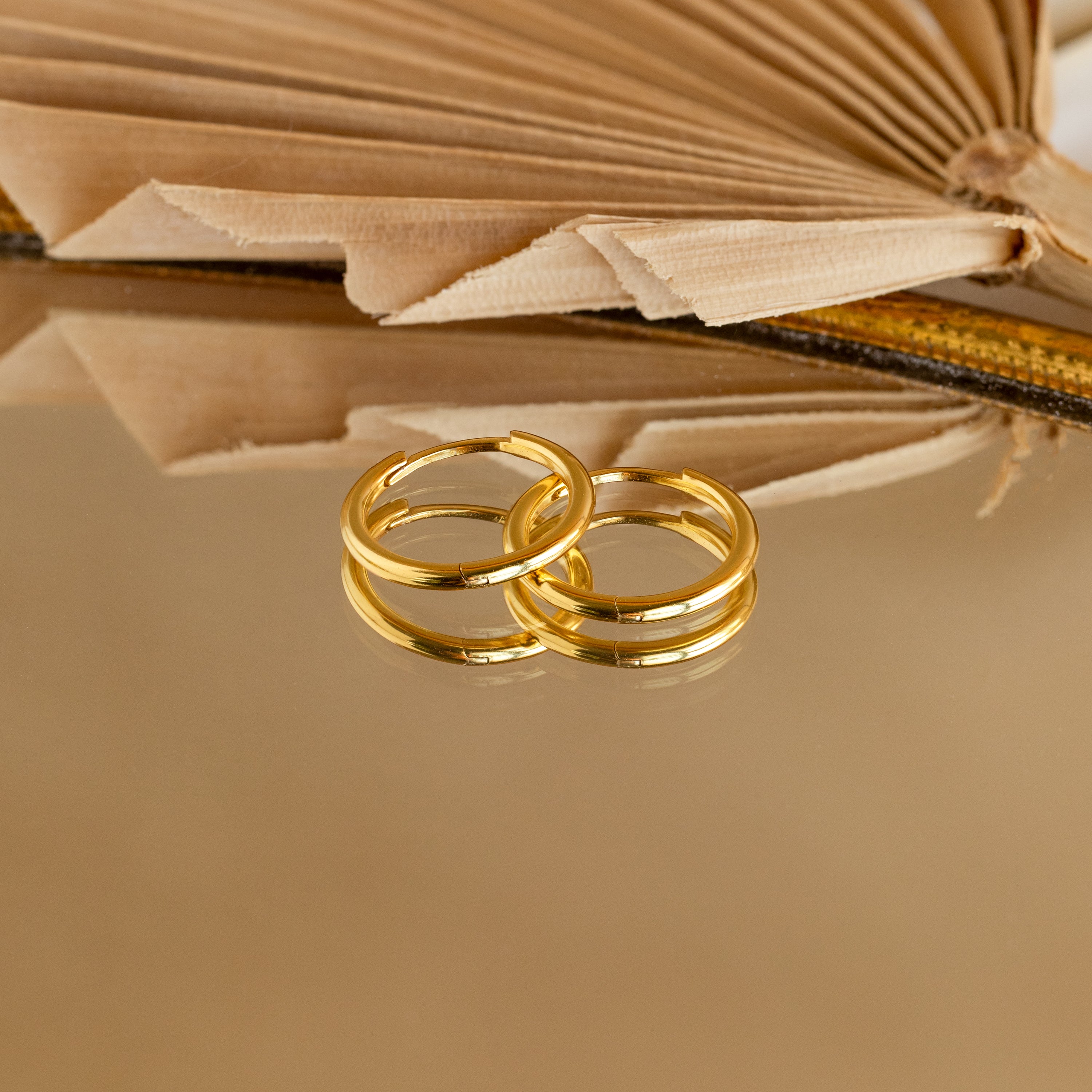 Two lightweight Medium Bold Hoops in 18K Gold rest on a reflective surface with an open book behind them. Perfect for everyday wear, these earrings bring a touch of elegance to your daily look.