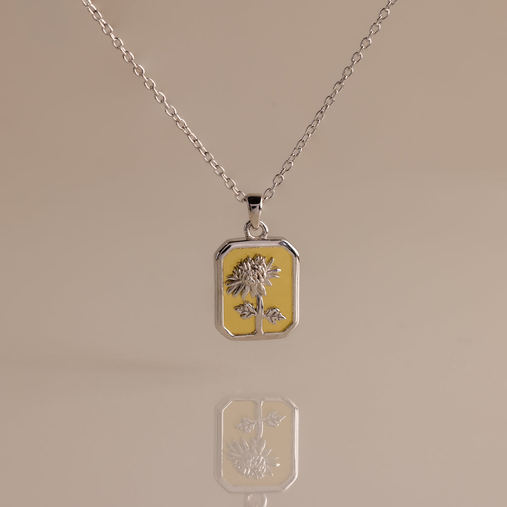 The Topaz Birth Flower Charm Necklace features a rectangular sunflower pendant, beautifully displayed on a beige background with an elegant reflection—ideal for those seeking a unique and delicate jewelry statement.