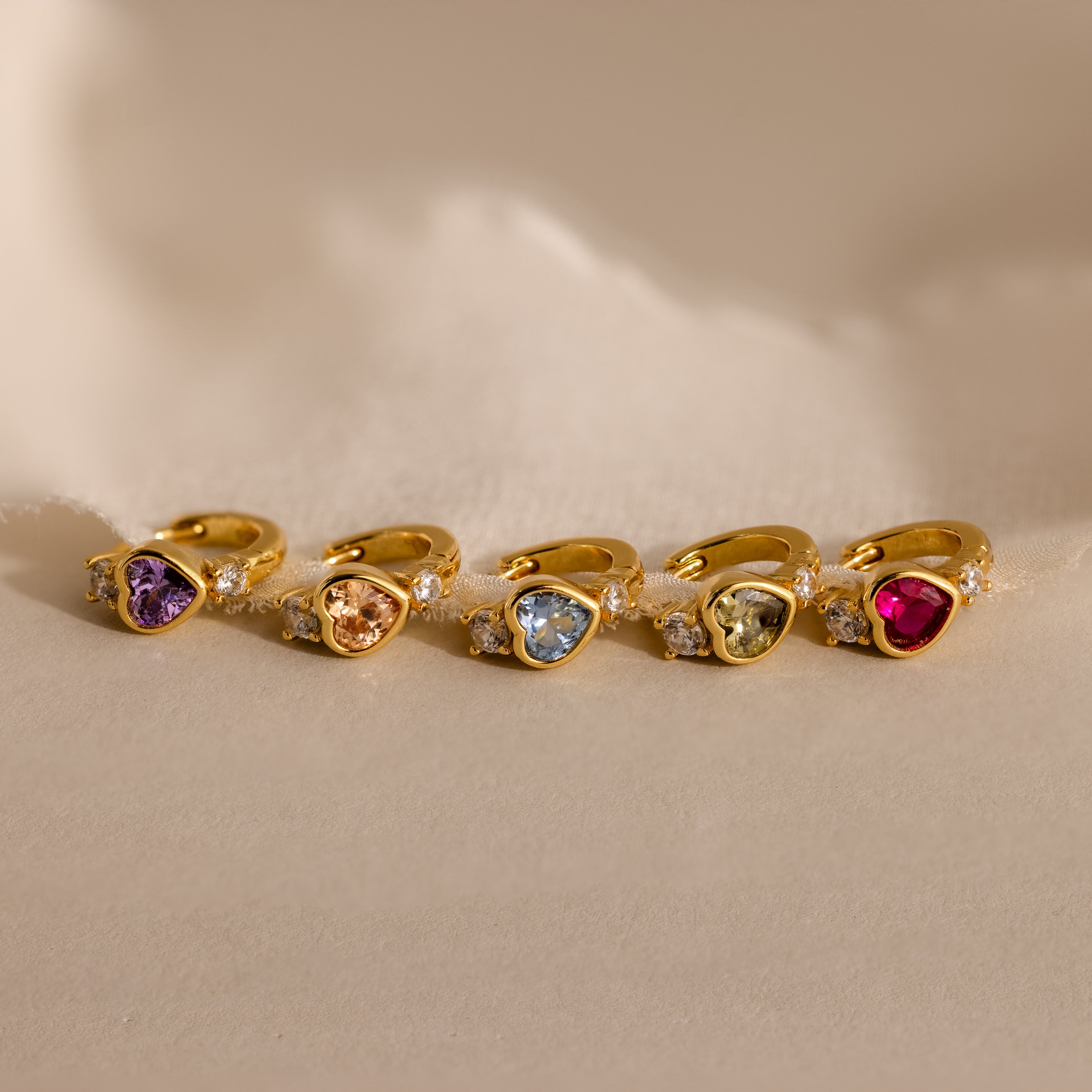 Five pairs of Birthstone Heart Hoops in different gemstone colors laid flat on their side