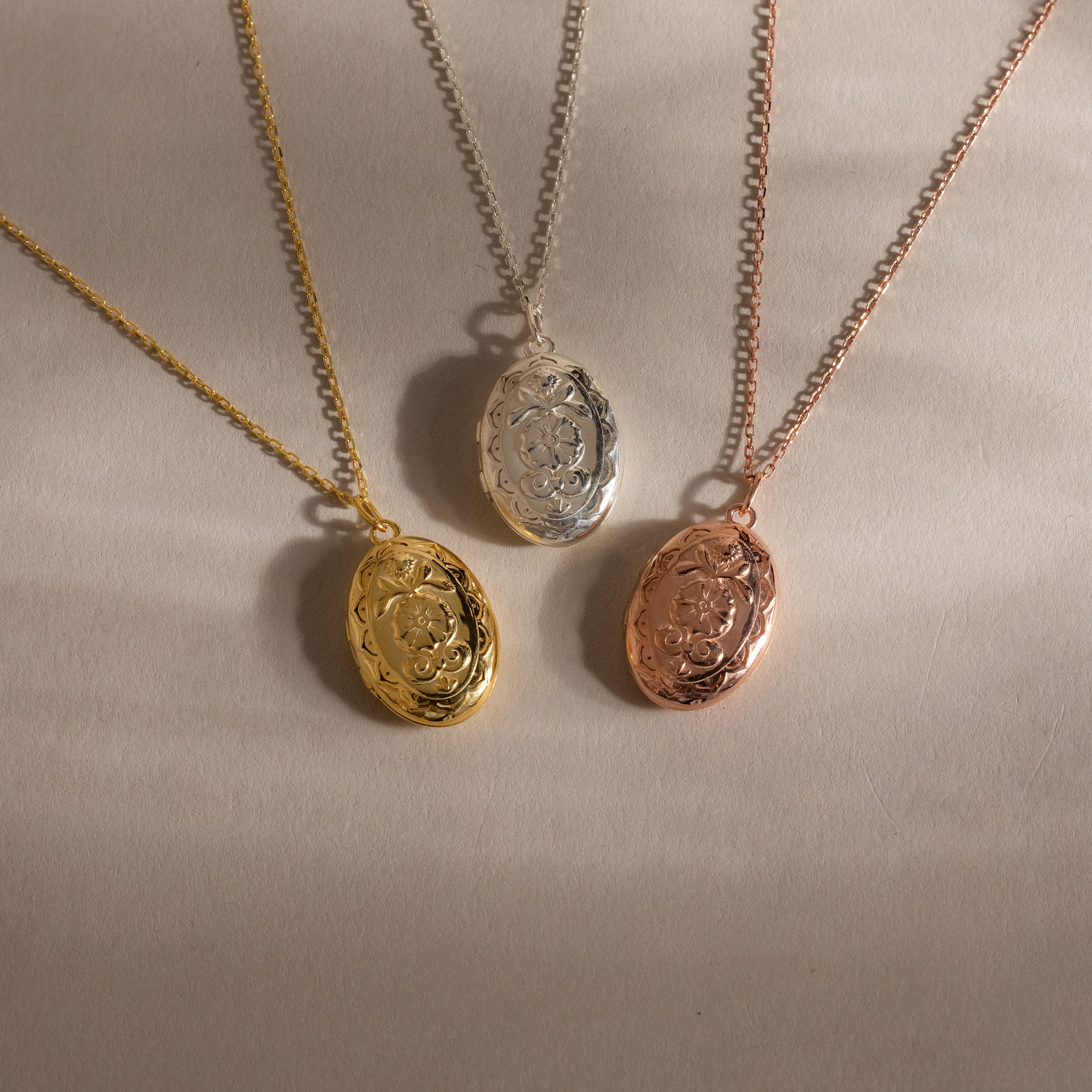 The Floral Handwriting Locket Necklace comes in gold, silver, or rose gold with a matching chain. Each oval locket can be personalized with your handwriting engraving, making it a meaningful keepsake. Displayed on a beige background.