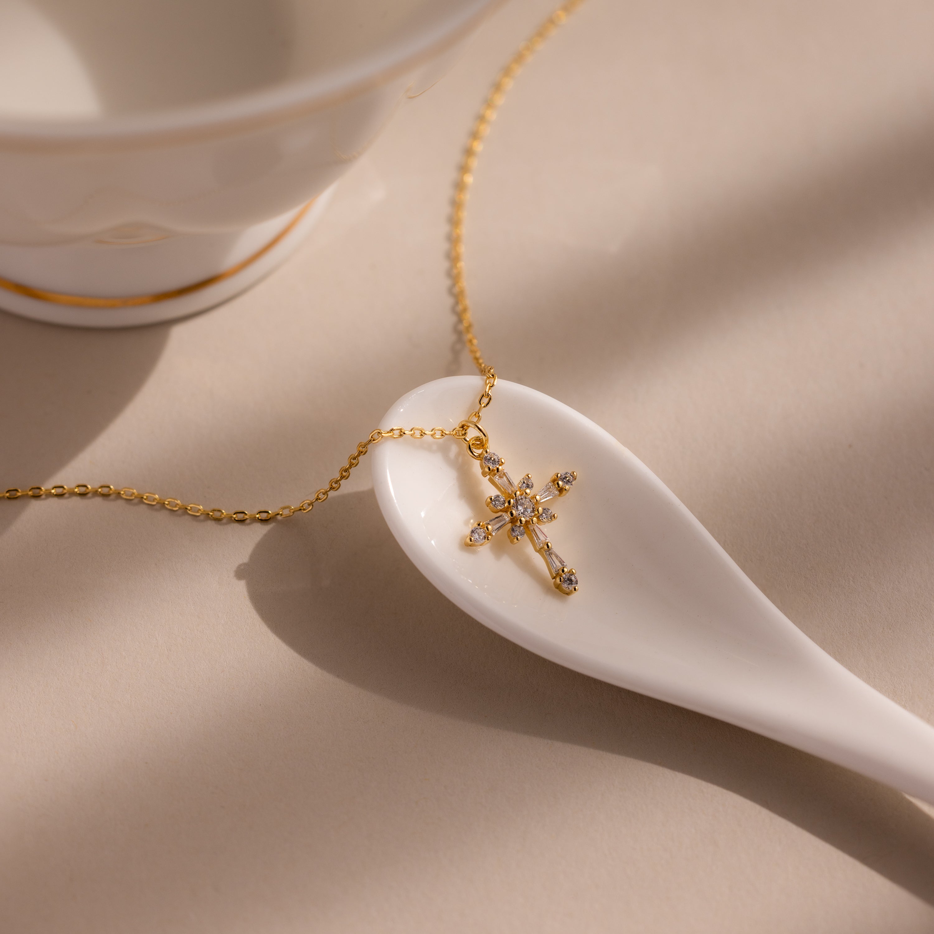 A Vintage Diamond Cross Necklace in 18K Gold rests on a white ceramic spoon beside a teacup.