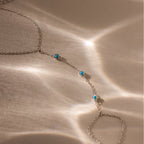 The Turquoise Hand Chain Bracelet in Silver features three petite turquoise stones, showcased on a softly lit beige surface.