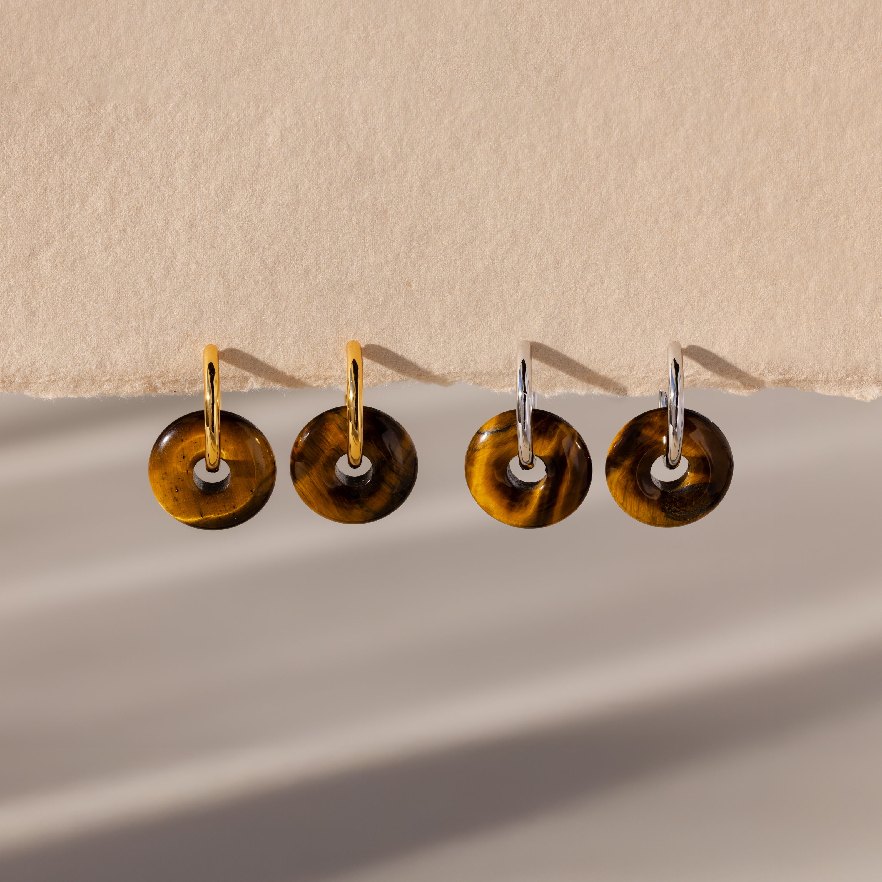 The Tiger's Eye Duo Hoops, featuring round tiger's eye gems with gold and silver hoops, are displayed on a beige textured surface and cast soft shadows.