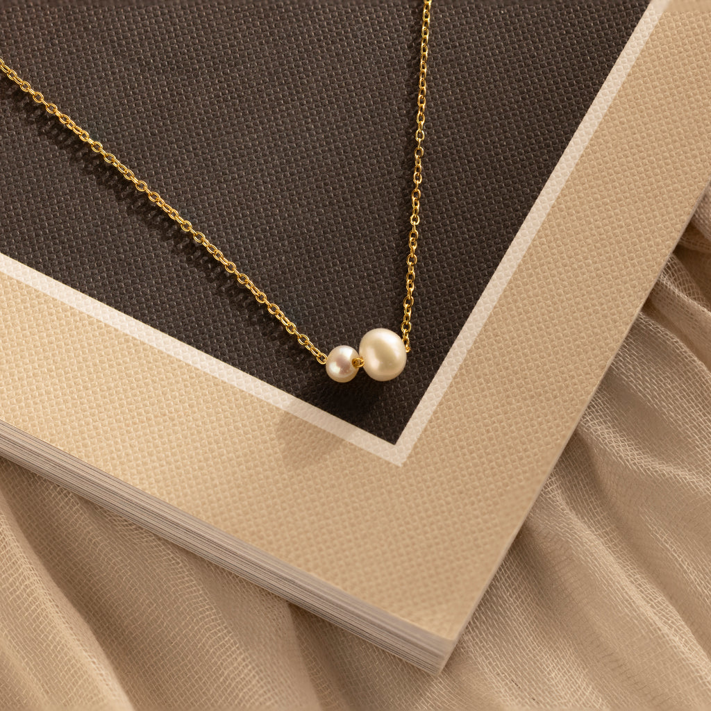 The Dainty Duo Pearl Necklace in 18K Gold, featuring two pearls on a textured beige and brown background, is perfect as wedding jewelry or a thoughtful gift for someone special.