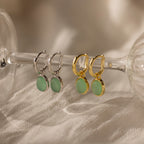 Two pairs of Jade Drop Hoops—one silver, one gold—with green jade stones displayed on a glass stem. Wear as elegant huggies or showcase their classic jade design.