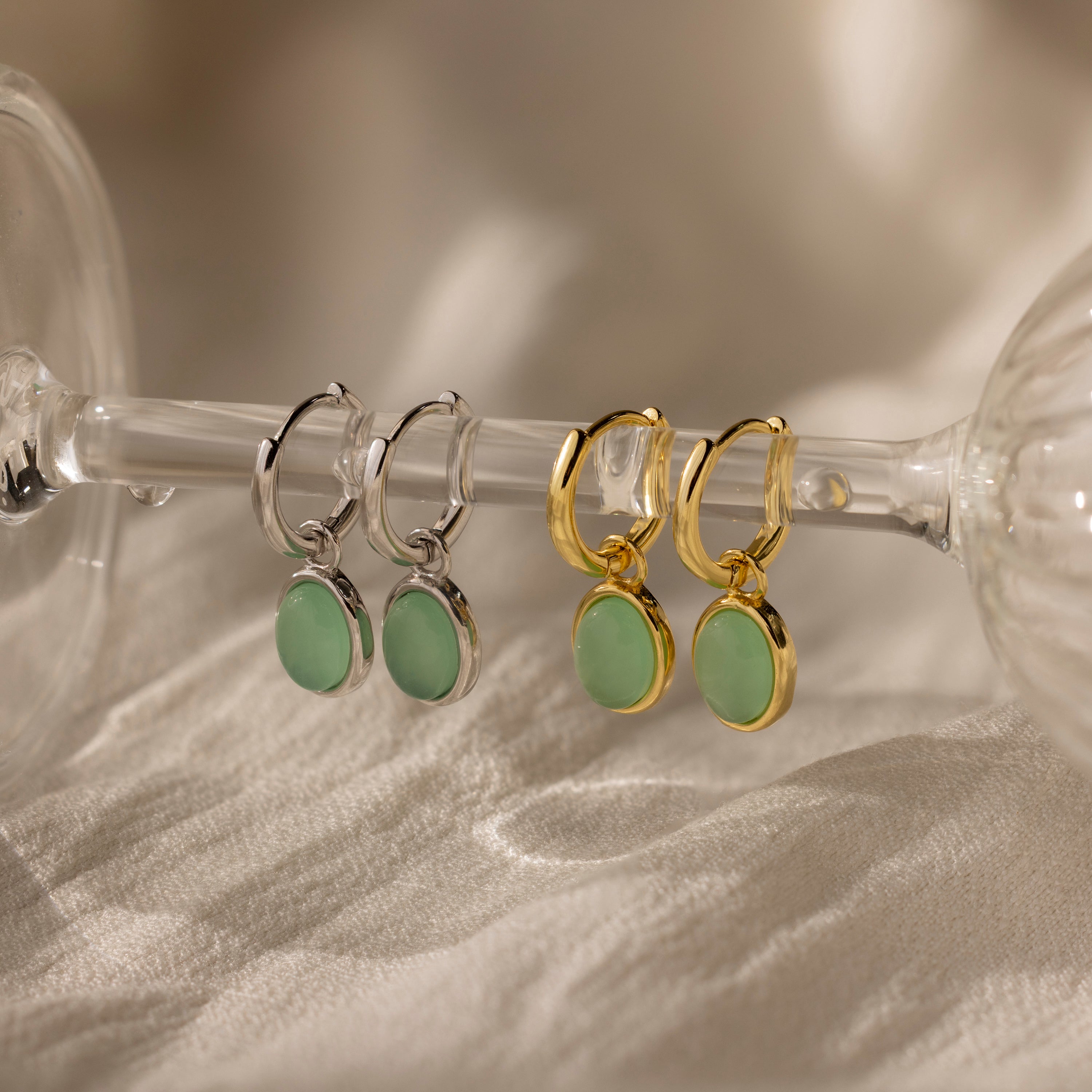 Two pairs of Jade Drop Hoops—one silver, one gold—with green jade stones displayed on a glass stem. Wear as elegant huggies or showcase their classic jade design.