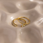 Two delicate gold rings from the Baguette Opal Ring Set, each adorned with a petite opal, rest on a beige, wavy-textured surface—an elegant set perfect for stacking or wearing alone.
