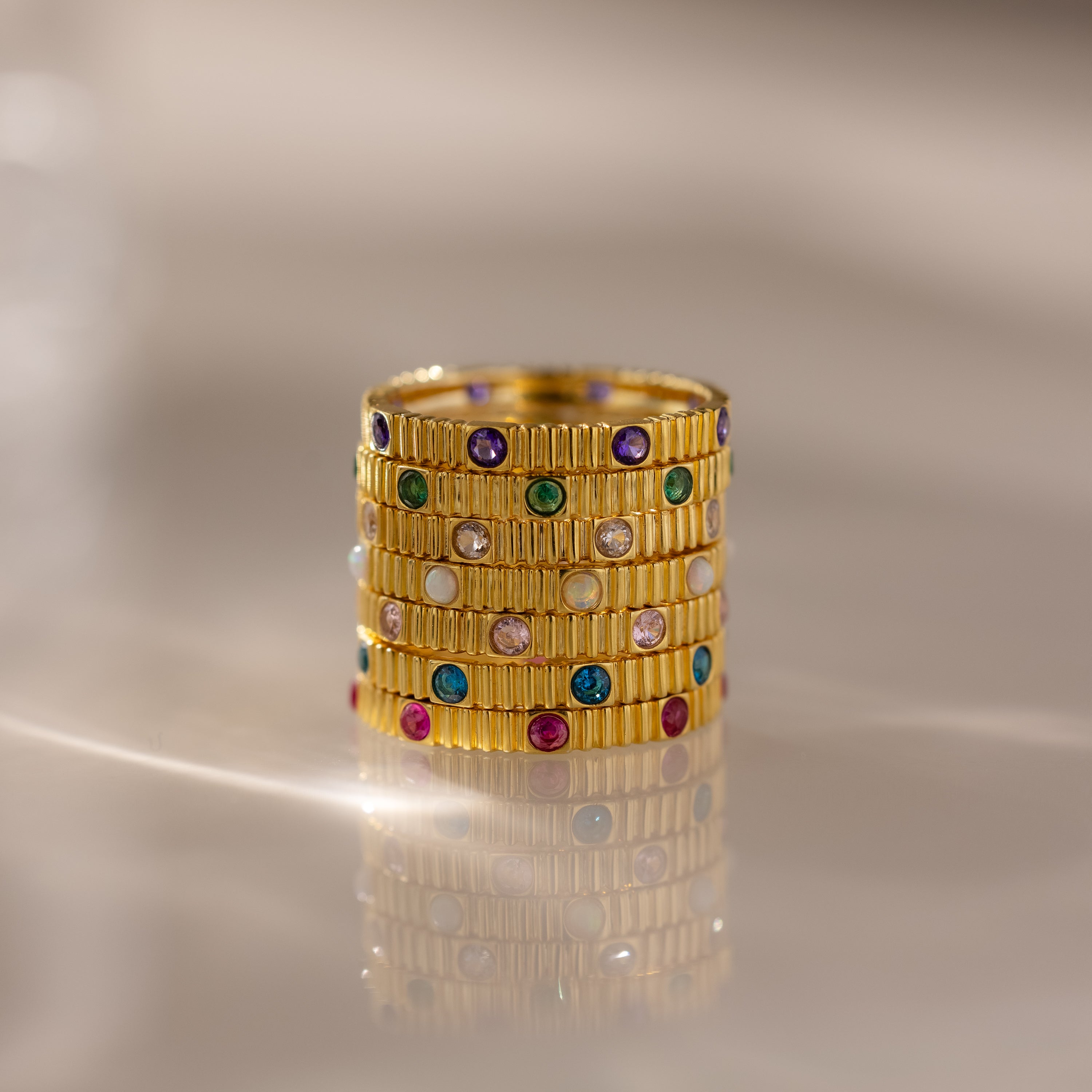 A Ribbed Round Birthstone Eternity Ring stacked with other gold rings, featuring colorful gemstones, displayed on a reflective surface.