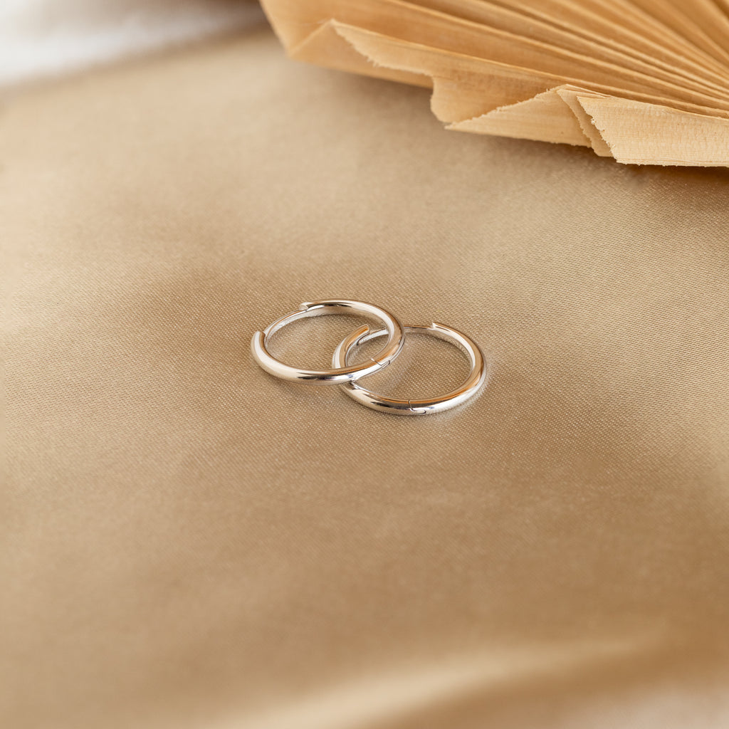 The Medium Bold Hoops in Sterling Silver rest on a beige satin surface with a book in the background, making them perfect for everyday wear.
