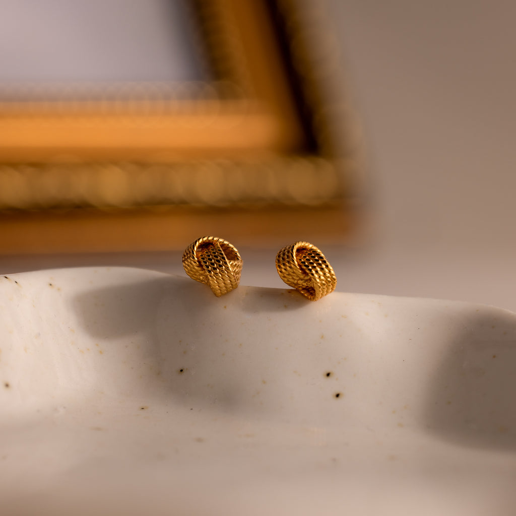 Rope Knot Studs in 18K Gold are displayed on a white textured surface with a blurred gold frame behind them, offering an elegant and timeless jewelry statement.